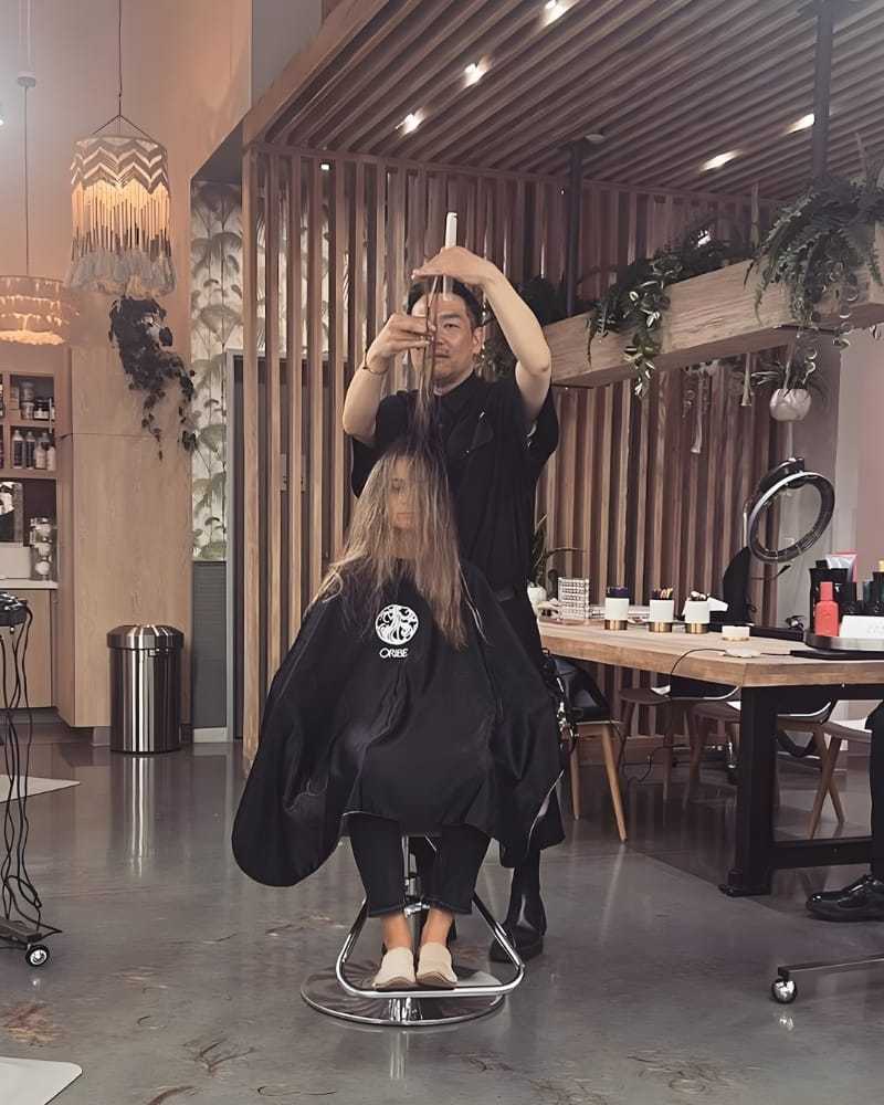 Hairstylist cuts a client's hair in a modern salon with wood accents and greenery.