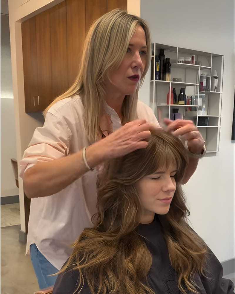 Hairstylist styles a woman's hair in a salon with shelves of products in the background.